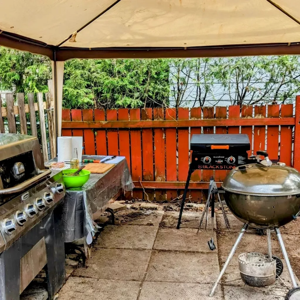 A gas grill, a Blackstone Griddle, a Weber Kettle Grill and a prep table under a canopy