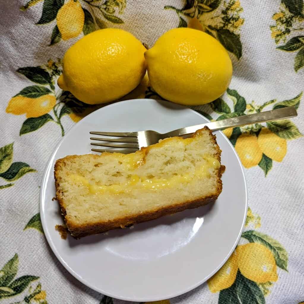 slice of lemon curd cake on a white plate with two lemons resting on a lemon patterned cloth