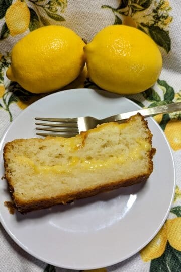 slice of lemon curd cake on a white plate with two lemons resting on a lemon patterned cloth