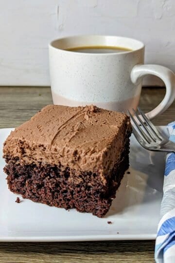 square of chocolate cake on a white plate with a fork and blue napkin. a cup of coffee in the background