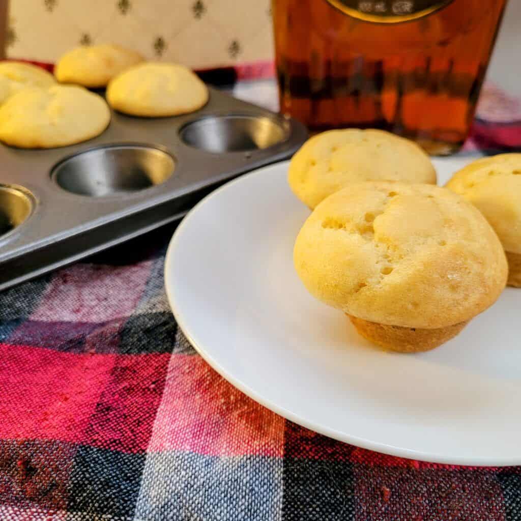 three pancake muffins on a plate with a bottle of maple syrup and a muffin pan in the background