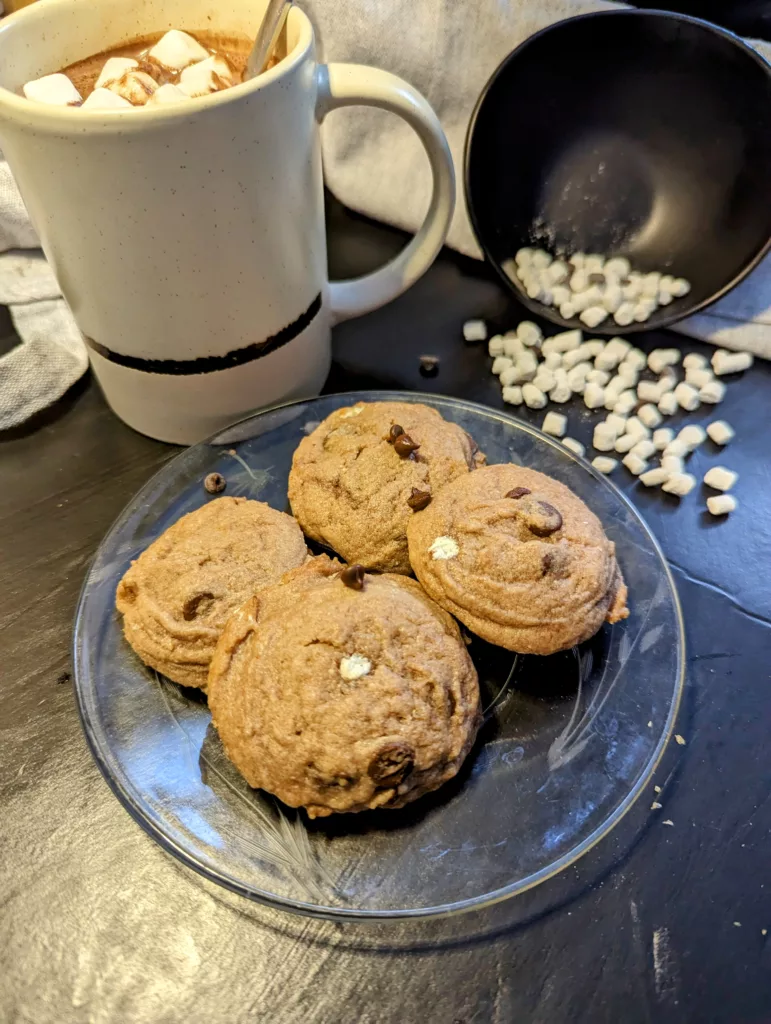 hot chocolate cookies on a plate with a cup of hot chocolate with marshmallows and a bowl of mini marshmallows spilled in the backround