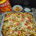 Pan of Fritos Chicken Casserole topped with pickled jalapenos and diced tomatoes. A bag of BBQ Fritos and a bowl of sour cream in the background