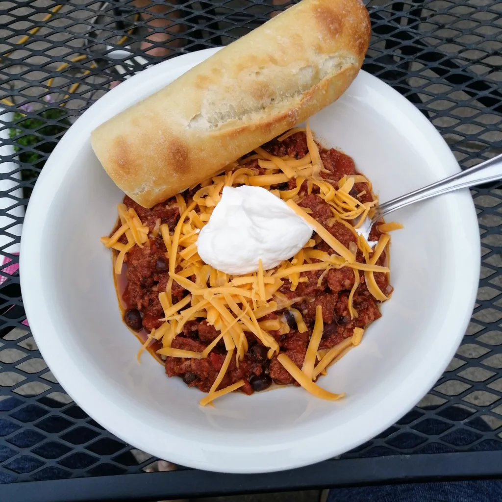 Bowl of chili topped with a dollop of sour cream and shredded cheese with a half baguette resting on the side of the bowl