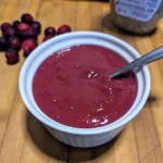bowl of cranberry mustard dipping sauce with a spoon in it and Dijon mustard and cranberries in the background on a wooden cutting board.
