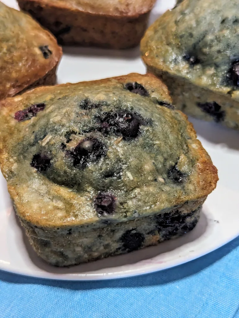Close up of square blueberry oatmeal muffin on a white plate with the edges of three more square blueberry muffins in the background.