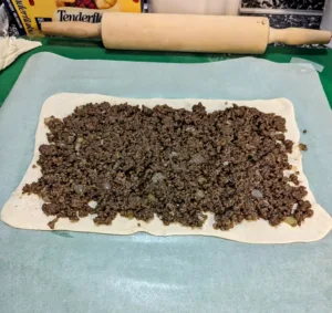 tourtiere meat on a sheet of puff pastry on parchment paper with a rolling pin in the background