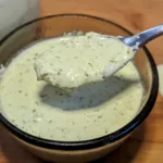A small bowl of creamy cilantro jalapeno sauce with a spoon of sauce hovering above the bowl