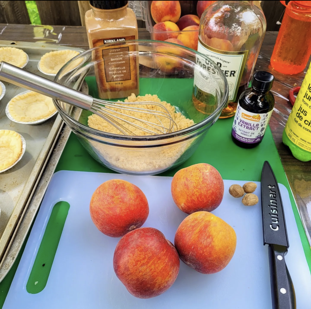 Four fresh peaches and whole nutmeg on a cutting board, with a knife beside them. There is a bottle of Bourbon, a bowl of brown sugar, a bottle of vanilla extract, a container of cinnamon and tart shells in the background.