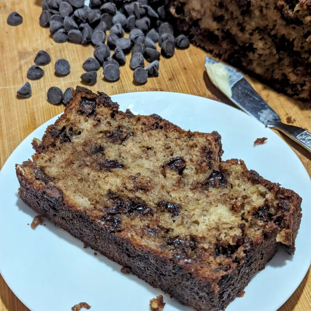 Slice of Chocolate Chip Banana Bread on a plate with chocolate chips spilled on the side and a butter knife with a small amount of butter