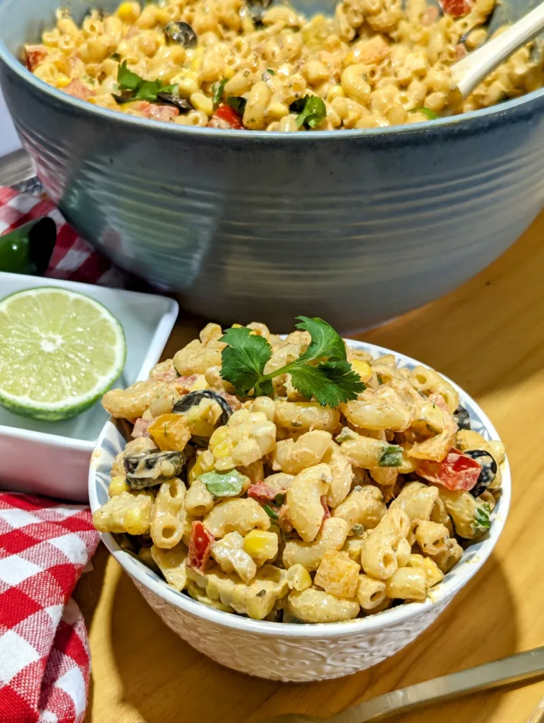 A small bowl of tex-mex macaroni salad with a large bowl of salad and a lime in the background.