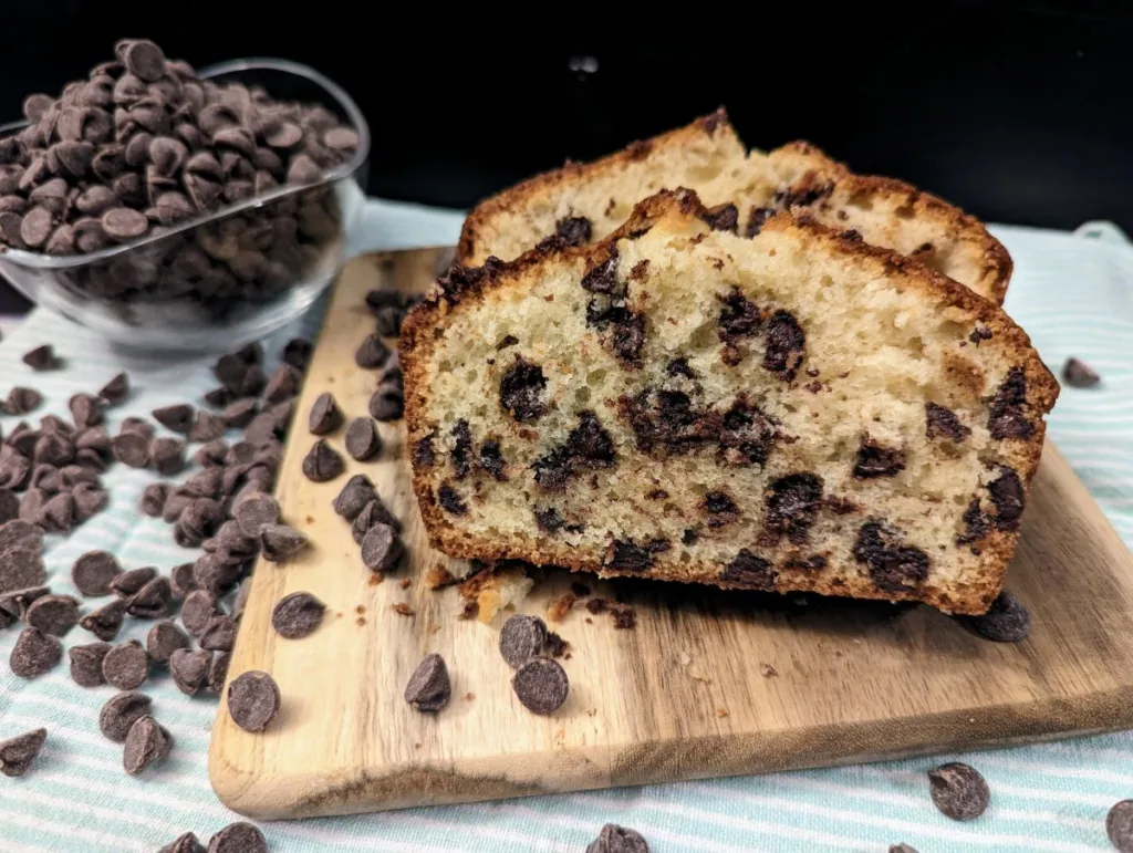 Sliced chocolate chip bread on a wooden cutting board with a clear bowl of chocolate chips in the back ground.
