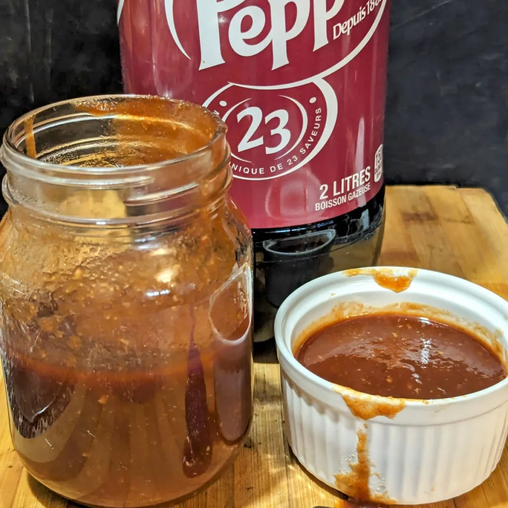 A mason jar half filled with Dr Pepper Rib Sauce beside a ramekin of sauce and a bottle of Dr Pepper in the back.